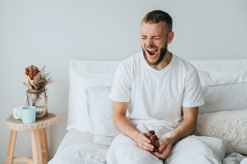 Sleepy young man in bed yawns holds jar with sleeping pills, feels fatigue, needs healthy sleep